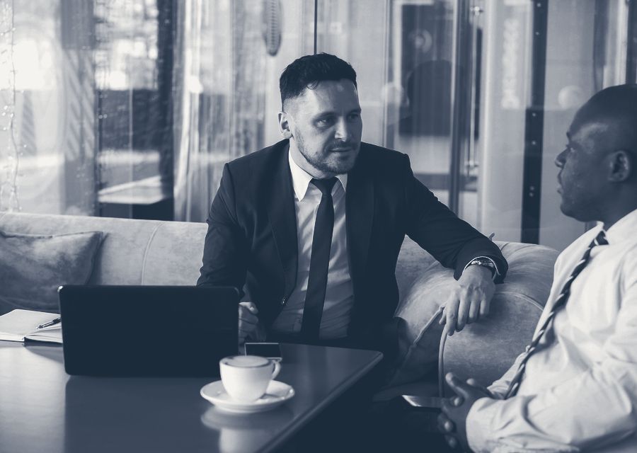 Two professionals in a glass-walled office, in conversation over coffee